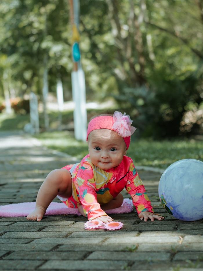 Adorable baby girl in bright outfit crawling outdoors on a sunny day beside a ball.