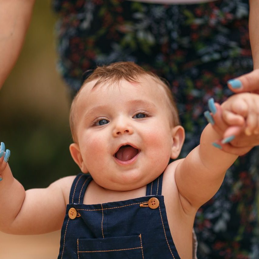 Cute baby smiling while learning to walk outdoors, holding parent's hands.