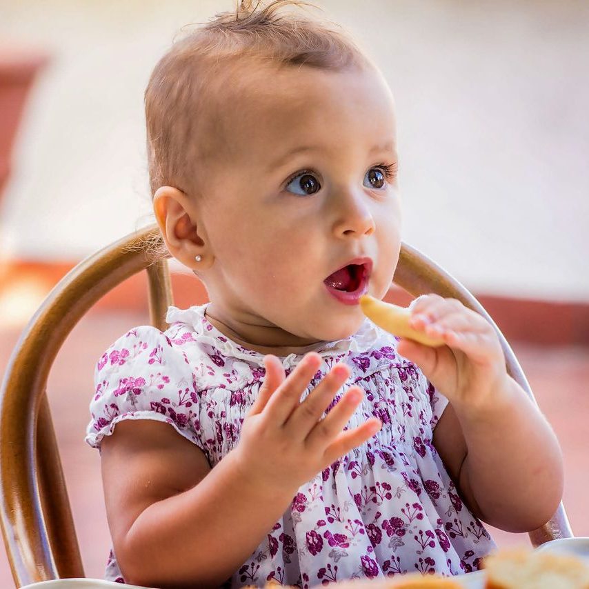 Cute baby girl in floral dress munching on a snack, enjoying a sunny day outside.