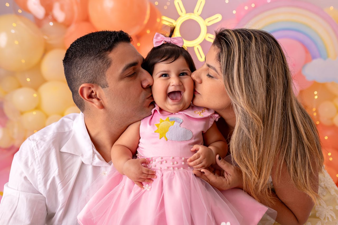 Joyful family portrait with parents kissing their laughing baby in colorful setting.