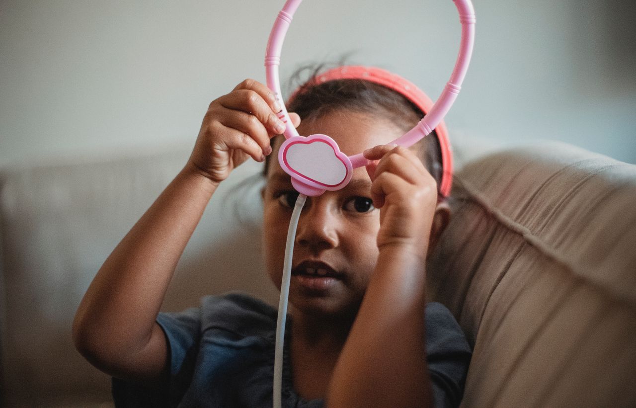 Pretty ethnic little girl looking at camera while sitting on couch with raised arms and pink toy stethoscope in hands