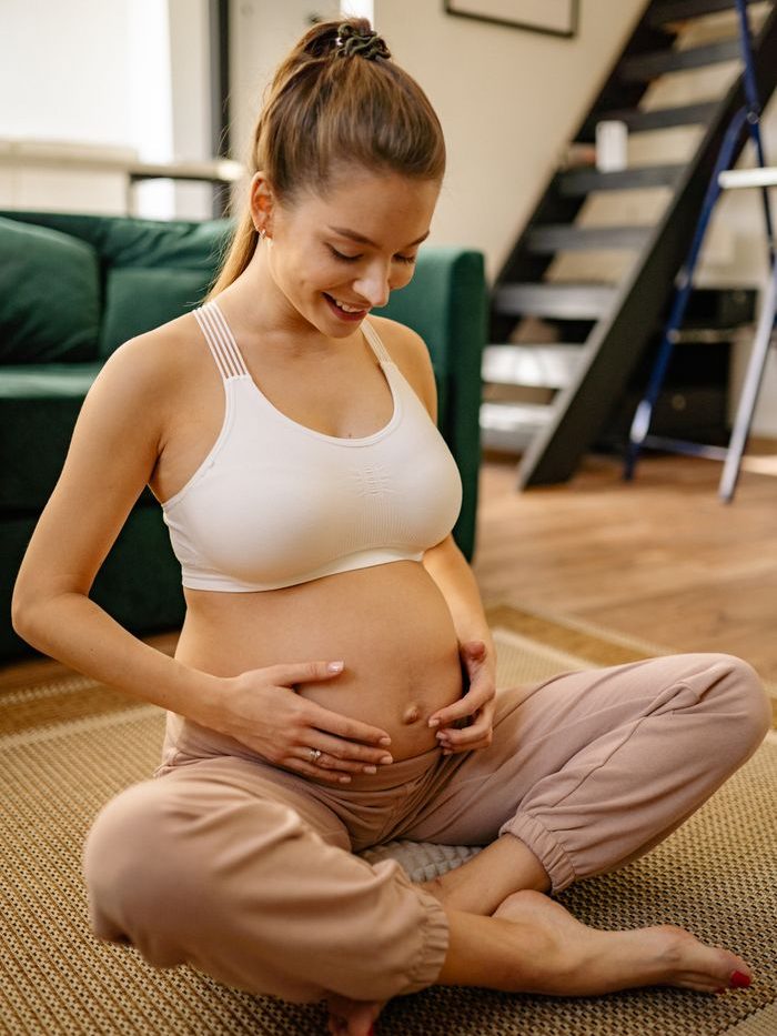 A smiling pregnant woman sits indoors, embracing her belly with love.
